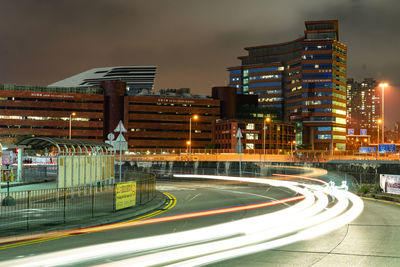 Light trails on road by buildings against sky at night