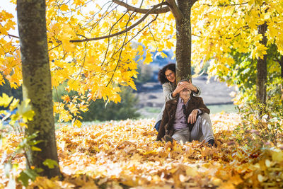  a man sits on the ground, a woman covers his eyes with her hands. mature couple in an autumn park. 