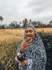 Portrait of senior woman smoking cigarette while standing at farm against sky