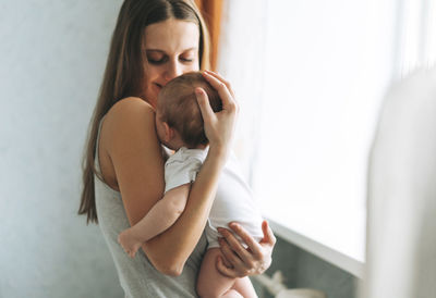 Young woman mom with long hair holding baby girl on hands near window at home