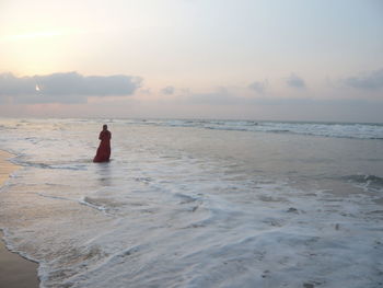 Rear view of woman standing on shore at puri beach against sky during sunset