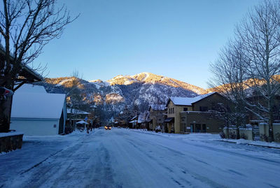 Snow covered road by trees and buildings against sky