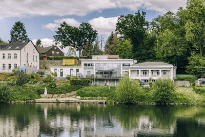 Trees and houses by lake against sky