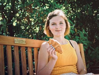 Portrait of smiling woman eating food while sitting on bench against trees