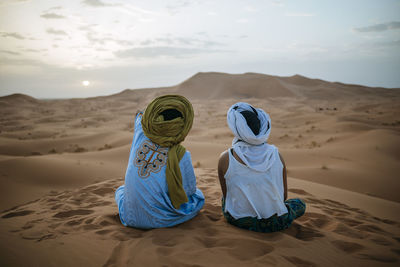 Woman sitting in the desert with berber guide, watching sunset