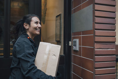 Smiling female messenger looking away while carrying box by closed door