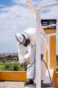 Anonymous employee in protective uniform and mask using air brush to paint frame structure against cloudy sky on sunny day