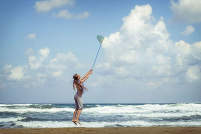 Girl holding fishing net while jumping at beach against sky