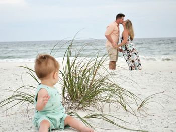 Toddler sitting on sea shore while parents kissing in background