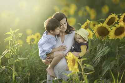 Woman is standing in the middle of a sunflower field with her son and her daughter in her arms