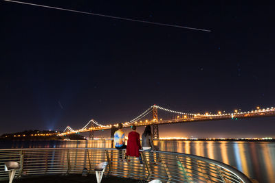 Rear view of people sitting on railing against illuminated bay bridge at night