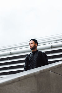 Young man looking away while standing by railing against sky