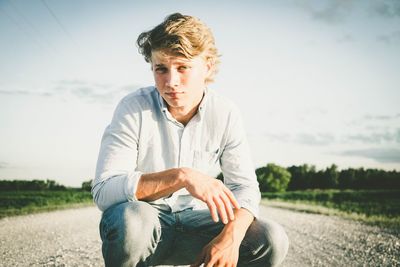 Portrait of young man crouching on street against sky