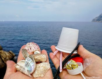 Cropped hands holding garbage and seashells at sea against sky