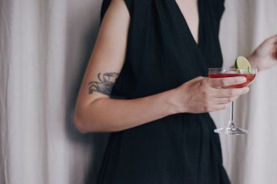 Midsection of young woman holding drink while standing curtain at home