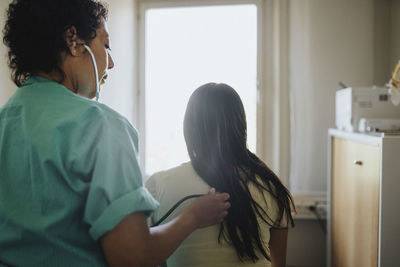 Rear view of mature female doctor examining young woman with stethoscope in clinic