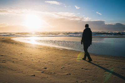 Rear view of man walking on beach against sky during sunset