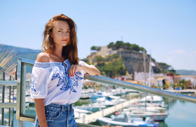 Portrait of young woman standing by railing against sky