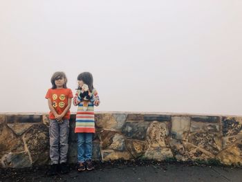 Children standing against stone wall against sky