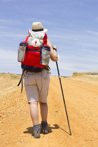 Rear view of hiker in hat with backpack walking on dirt road during sunny day