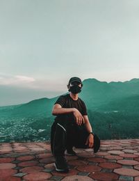 Young man sitting on mountain against sky