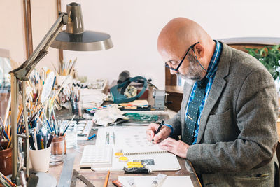 Side view of focused creative mature male designer working on jewelry designs and drawing sketches while sitting at table with professional supplies in workplace