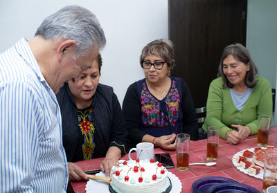 An older man with his friends celebrating a birthday, cutting a cake at an indoor party.