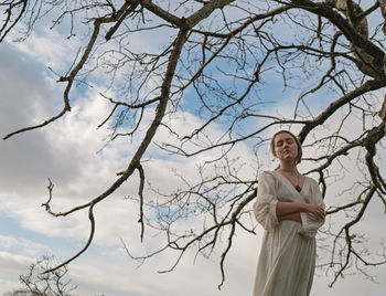 Low angle view of woman standing by bare tree against sky