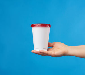 Female hand holds a disposable white glass with a red cap on a blue background, take-away drinks