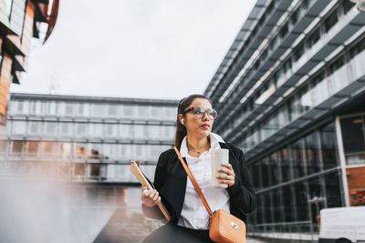 Businesswoman looking away holding insulated drink container while standing in front of building
