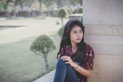 Beautiful young woman looking away while sitting against wall