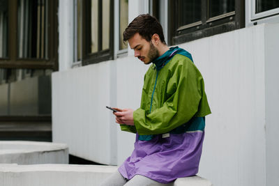 Man using mobile phone while sitting on retaining wall against building