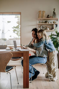 Son embracing mother working while sitting near dining table at home