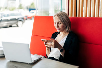 A self-employed woman is working with her phone and laptop on the terrace of a restaurant over a cup