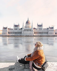 Woman in warm clothes sitting by river against historic building 