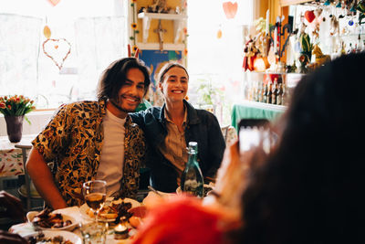 Young woman photographing smiling friends sitting at table in restaurant during brunch