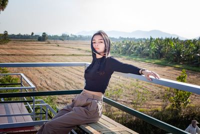 Young woman sitting on railing against sky