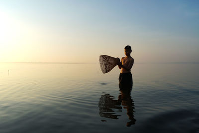 Full length of shirtless man holding sea against sky during sunset