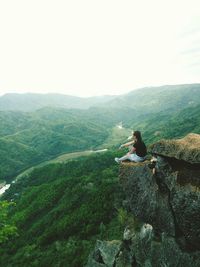 Man sitting on rock looking at mountain against sky