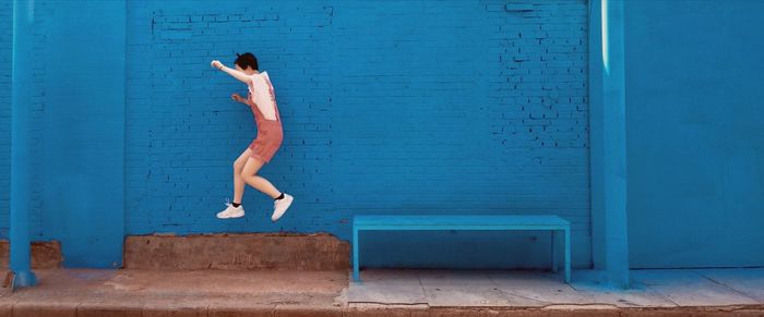 Side view of young woman jumping by blue wall