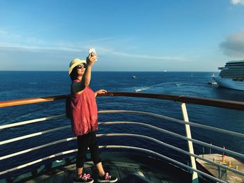 Woman taking selfie on mobile phone while standing by railing against sea and sky