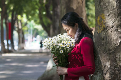 Woman smelling flowers in bouquet while standing by tree trunk