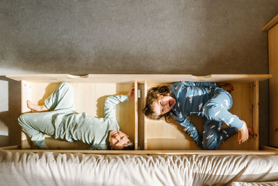 Top view of happy siblings in nightwear looking at camera while lying in wooden drawers of bed in light bedroom