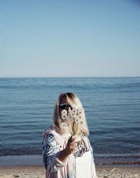 Mid adult woman holding dandelion seeds while standing at beach against clear sky