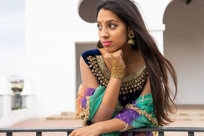 Positive young ethnic brunette in sari with makeup and accessories standing on balcony of white building and leaning on railing while looking away