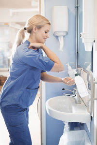 Side view of young nurse using soap dispenser to wash hands in bathroom