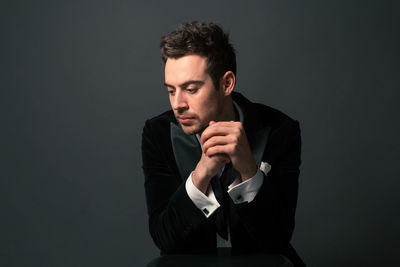 Thoughtful young man in suit sitting with hands clasped against gray background
