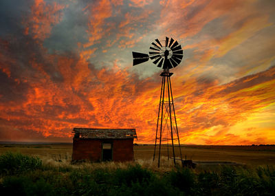 Water pump windmill by abandoned barn on field against sky at sunset