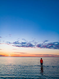 Rear view of boy standing in sea against sky during sunset