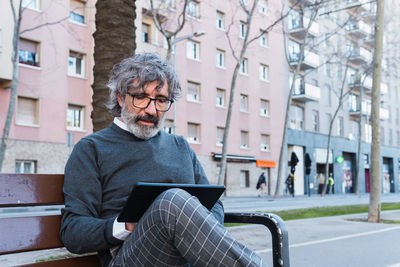 Handsome mature man in eyeglasses using digital tablet on bench in the street